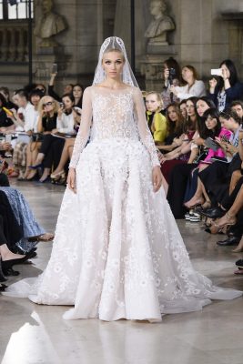 Georges Hobeika’s model wears an elaborate wedding gown, with a fragile bodice and sheer sleeves, hand-sewn crystals and a theatrical veil cascading from her crown. It took four members of the Atelier staff to carry the veil and train onto the catwalk.