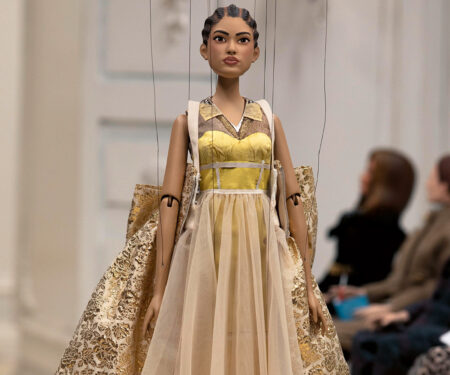 A doll walks the Moschino runway in place of models, wearing a gold dress