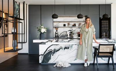 Sally Matthews, wearing a khaki coat, stands in front of her marble kitchen island.