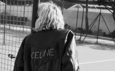 Model sits on fence wearing Celine Homme Summer '22 black leather jacket