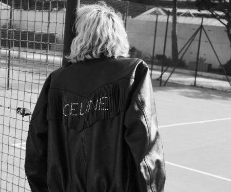 Model sits on fence wearing Celine Homme Summer '22 black leather jacket
